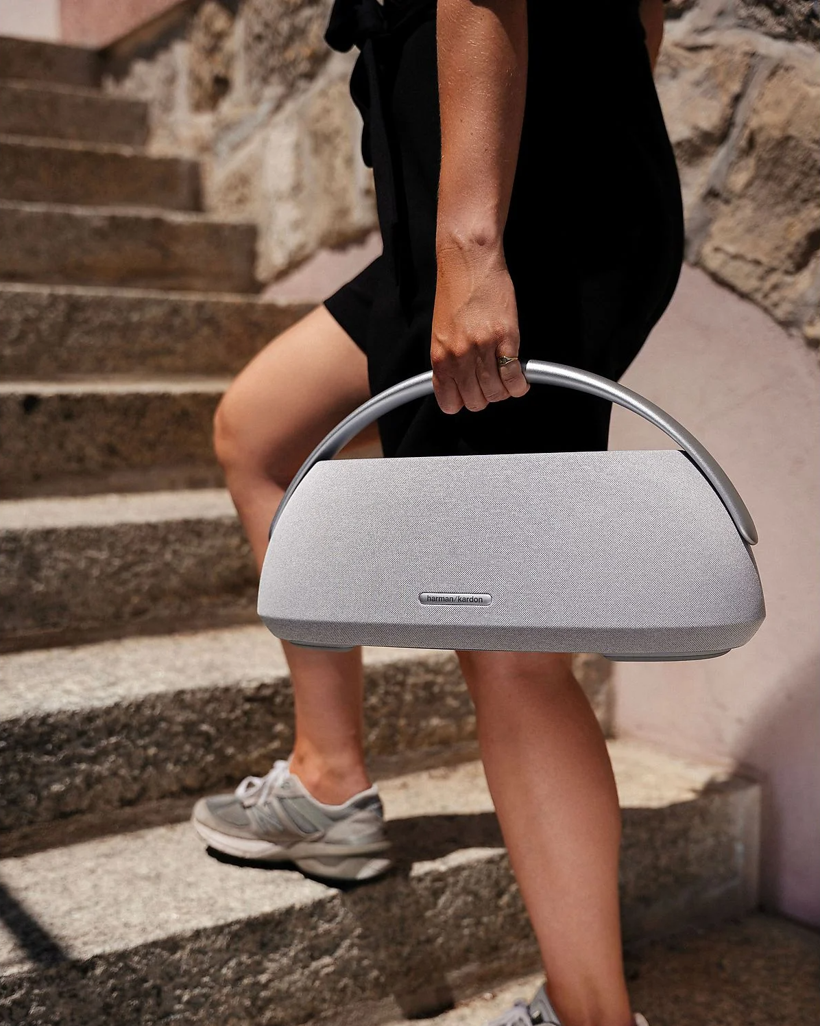Harman Kardon GOPLAY3 Bluetooth Hoparlör Gri, dokulu kumaş ve metal tutma sapıyla merdivenlerde taşınıyor.