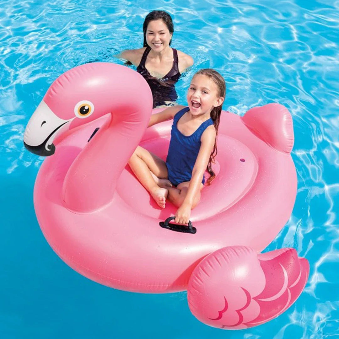 Mavi havuzda küçük bir kızın üzerinde oturduğu pembe renkli Intex flamingo deniz yatağı.