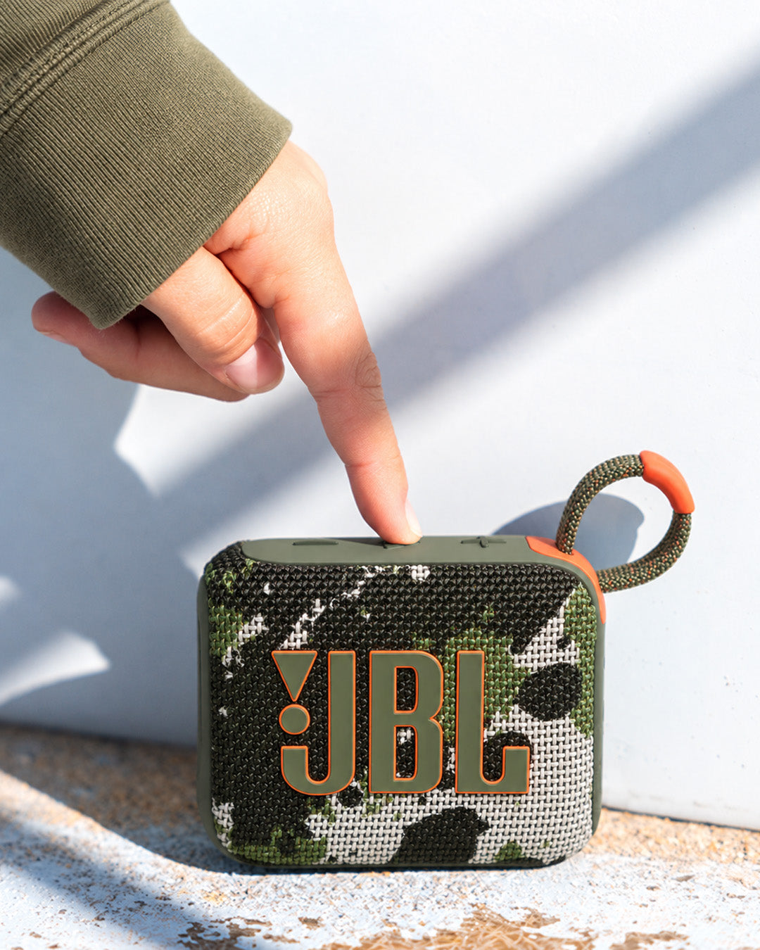 JBL Go 4 taşınabilir Bluetooth hoparlör, kamuflaj desenli gövdesi ve askı kancasıyla dış mekanda bir el tarafından açılıyor.