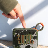 JBL Go 4 taşınabilir Bluetooth hoparlör, kamuflaj desenli gövdesi ve askı kancasıyla dış mekanda bir el tarafından açılıyor.