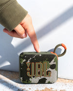 JBL Go 4 taşınabilir Bluetooth hoparlör, kamuflaj desenli gövdesi ve askı kancasıyla dış mekanda bir el tarafından açılıyor.