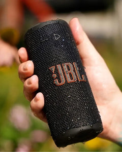 JBL Grip Taşınabilir Bluetooth Hoparlör siyah renkli, su damlacıklı dokulu ızgarası elde tutulurken.